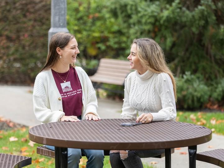 Two students sit outside at a table during a beautiful fall day